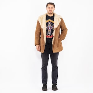 Könnte beinhalten: Ein brauner Wildledermantel mit cremefarbenem Shearling-Kragen und -Futter. Der Mantel hat einen Knopfverschluss und Taschen. Die Person trägt ein schwarzes T-Shirt mit Grafikdesign und dunkelblaue Hose.