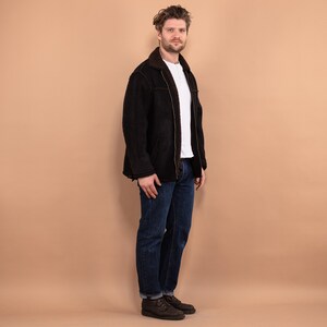 May include: A man wearing a black suede jacket with a brown shearling collar, blue jeans, and brown leather boots.