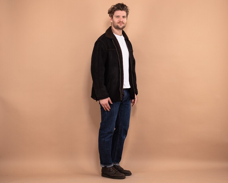 May include: A man wearing a black suede jacket with a brown shearling collar, blue jeans, and brown leather boots.