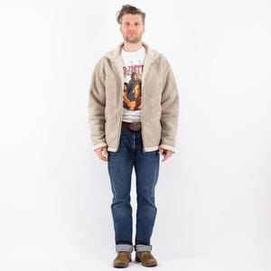 May include: A person wearing a tan shearling-lined jacket with a hood, over a white graphic t-shirt and blue jeans. The jacket has a zipper closure and a brown belt. The person is wearing brown shoes.