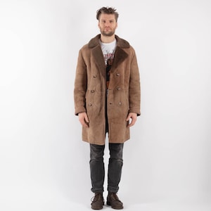 Puede incluir: Un abrigo marrón de doble botonadura con cuello y forro de borrego. El abrigo es largo y abotonado. La persona lleva una camiseta blanca y vaqueros azul oscuro. El abrigo es de estilo vintage.