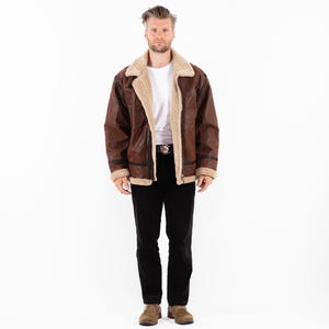 May include: A brown leather jacket with a cream-colored shearling lining and collar. The jacket has a zip closure and black accents on the sleeves. The person is wearing a white t-shirt and black pants.