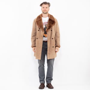 Puede incluir: Un abrigo cruzado color beige con cuello y puños de borrego marrón. El abrigo tiene botones marrones oscuros y un diseño clásico. La persona lleva una camiseta blanca y vaqueros grises. El abrigo es de estilo vintage.