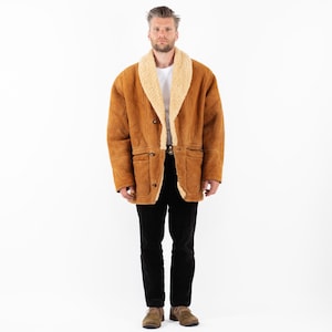 Puede incluir: Un abrigo de ante marrón con cuello y forro de borrego color crema. El abrigo tiene dos bolsillos delanteros y cierre de botones. La persona de la imagen lleva una camiseta blanca, pantalones negros y zapatos marrones.