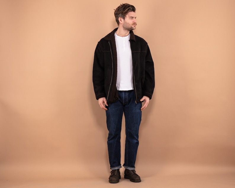 May include: A man wearing a black suede jacket with a white t-shirt underneath. He is wearing blue jeans and brown boots. The man is standing in front of a light brown background.