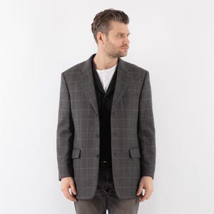 Puede incluir: Una chaqueta gris a cuadros con un chaleco negro debajo. La chaqueta tiene cierre de botones y dos bolsillos en la parte delantera.