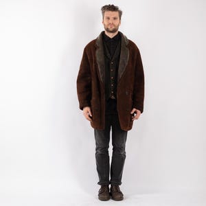 May include: A brown, double-breasted coat with a green collar and lapel. The coat is worn over a black shirt and vest, paired with dark gray jeans and brown shoes. The coat appears to be made of a textured material.