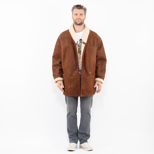 Puede incluir: Un abrigo de borrego marrón con forro y ribete de borrego color crema. El abrigo tiene una parte delantera abotonada, dos bolsillos delanteros y un estilo clásico y vintage. El modelo lleva una camiseta blanca y vaqueros grises.