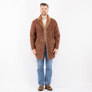 May include: A brown suede shearling coat with a fur collar and a button closure. The coat is worn over a white t-shirt and blue jeans.
