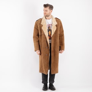 Puede incluir: Abrigo largo de ante marrón con cuello y forro de borrego beige. El abrigo tiene una botonadura frontal y charreteras. El modelo lleva una camiseta blanca con estampado gráfico y vaqueros negros.