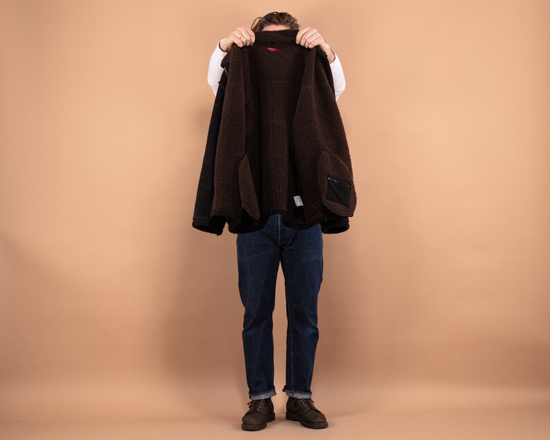 May include: A brown fleece-lined jacket with a black outer layer. The jacket is being held open by a person, revealing the fleece lining.