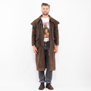Könnte beinhalten: Ein brauner, langer Duster-Mantel mit breitem Kragen und Schulter-Capes, getragen über einem weißen Grafik-T-Shirt mit dem Schriftzug Led Zeppelin, einer dunkelgrauen Jeans und braunen Lederstiefeln. Der Mantel hat große Taschen und Knöpfe.