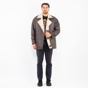 Puede incluir: Un abrigo de borrego marrón con forro y cuello de borrego color crema. El abrigo tiene un diseño cruzado con botones grandes y un estilo clásico y vintage. La persona de la imagen lleva pantalones grises oscuros y zapatos negros.