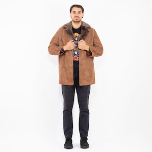 May include: A brown suede coat with a faux fur collar. The coat is buttoned and worn over a black graphic t-shirt and dark gray pants. The coat has a classic, vintage style.