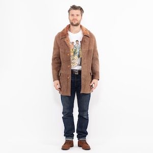 Puede incluir: Un abrigo de ante marrón con cuello y forro de borrego. El abrigo tiene cierre de botones, dos bolsillos delanteros y un cinturón. La persona lleva una camiseta blanca, vaqueros azules y botas marrones.