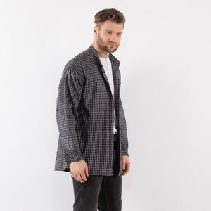 以下が含まれることがあります： 白と黒のチェック柄の長袖シャツに白いインナーを着た男性。シャツはボタンダウンカラーでゆったりとしたシルエットです。