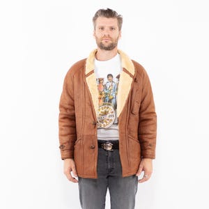 Puede incluir: Chaqueta de cuero marrón con forro y cuello de borreguillo color crema. La chaqueta tiene dos bolsillos delanteros y botones. La persona lleva una camiseta gráfica y vaqueros grises. El diseño de la chaqueta es clásico.