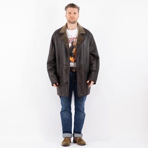 Puede incluir: Un abrigo de cuero marrón oscuro con cuello de borrego y cierre de botones. El abrigo se lleva sobre una camiseta gráfica blanca y vaqueros azules. El abrigo tiene un estilo clásico y vintage.