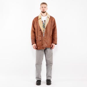 May include: A brown leather jacket with a tan shearling collar and lining. The jacket has a button-up front and is worn over a white t-shirt and gray pants. The person is wearing black shoes.