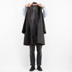 May include: A person wearing a dark gray coat with a black lining. The coat is open and the person is holding the lapels. The person is wearing dark blue jeans and black shoes.