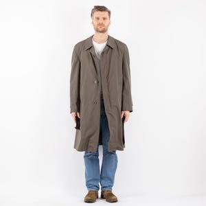 May include: A long, olive green trench coat with a button closure and a single button on each cuff. The coat has a single breast pocket and two side pockets. The model is wearing blue jeans and brown shoes.