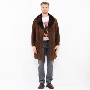 Puede incluir: Un abrigo de ante marrón con cuello de piel marrón oscuro. El abrigo tiene una botonadura frontal y se lleva sobre una camiseta blanca con gráficos y vaqueros grises. La persona lleva zapatos de cuero marrones.