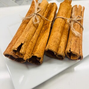 May include: Bundles of cinnamon sticks, tied with twine, rest on a white square plate. The cinnamon sticks are a warm brown color with a textured surface. The image is well-lit, highlighting the natural tones and textures of the spice.