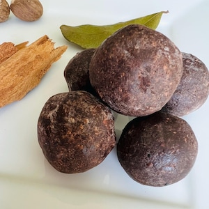 May include: A close-up of several dark brown, round spice balls, possibly truffles or similar, arranged on a white surface. Accompanying the spice balls are pieces of cinnamon bark, a bay leaf, and whole nutmeg.