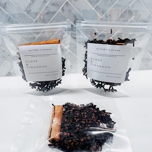 May include: Three clear plastic bags filled with spices. Two bags are upright, and one is open. Each bag contains cloves, cinnamon sticks, and a label that reads "Clove & Cinnamon" and "Nutmeg Nirvana". The spices are a dark brown color.
