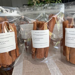 May include: Three clear plastic bags filled with cinnamon sticks. Each bag has a white label that reads "Nutmeg Nirvana" and "Cinnamon Sticks". The cinnamon sticks are a warm brown color and appear to be whole, dried pieces of cinnamon bark.