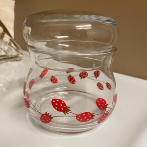 May include: A clear glass jar with a lid. The jar features a repeating pattern of red strawberries with white dots and green stems. The jar is designed for storing food items, such as jam or preserves.