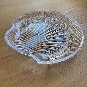May include: A clear glass serving dish with a scalloped edge and a textured, fan-like design. The dish has a shallow depth and is suitable for serving appetizers or small portions. The glass is transparent, allowing the contents to be seen.