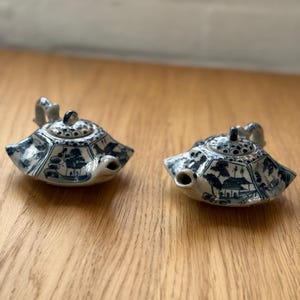 Puede incluir: Dos pequeñas teteras de cerámica azul y blanca con un diseño de paisaje detallado. Cada tetera tiene un pico, un asa y una tapa. Las teteras se exhiben sobre una superficie de madera de color marrón claro.