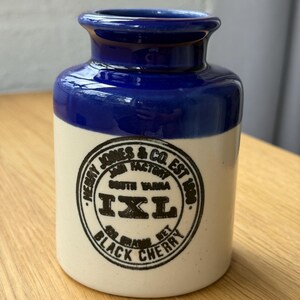 以下が含まれることがあります： 青い上部とクリーム色のベースの陶器の瓶。瓶には、「HENRY JONES & CO. JAM FACTORY EST 1898 SOUTH YARRA IXL BLACK CHERRY」というテキストが入った黒い円形のエンブレムが付いています。
