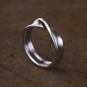 May include: A silver-colored ring with a twisted design. The ring has a smooth, polished finish and a rounded band. The twisted element creates a unique, eye-catching detail. The ring is shown on a wooden surface.