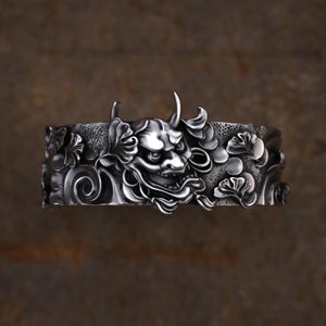May include: A silver bracelet with a detailed design of a Japanese demon face surrounded by stylized floral and wave patterns.