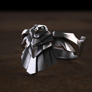 May include: A silver-colored ring featuring a stylized eagle design. The ring's focal point is the eagle's head and wings, with detailed feather patterns. The ring is made of a shiny metal and has a geometric, angular design.