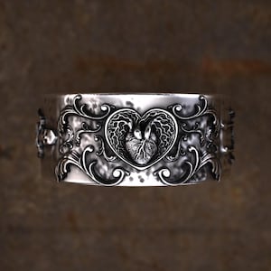 May include: Silver cuff bracelet featuring an intricate design. A heart-shaped emblem with a detailed anatomical heart is the focal point. The bracelet is adorned with swirling, floral patterns. The metal has a hammered texture.
