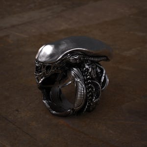 May include: A detailed, silver-colored ring featuring the head of an alien creature. The ring showcases intricate details of the alien's skull, head, and facial features. The ring is set against a dark, wooden background.