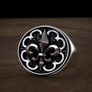 May include: A silver signet ring featuring a detailed fleur-de-lis design within a circular frame. The ring has a polished finish and a thick band. The design is centered on the ring's face, with intricate scrollwork around the edges.
