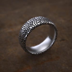 May include: A silver ring featuring a detailed, swirling pattern on the outer surface. The ring has a polished inner surface and a textured edge. The design is intricate and appears to be made of sterling silver.
