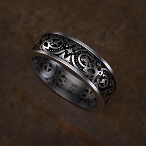 May include: A silver ring with an intricate, openwork design. The band features a repeating pattern of swirling shapes and floral elements. The ring is marked with "925" on the inside, indicating its silver content. The ring is displayed against a brown background.