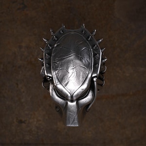 May include: A silver Predator mask with a detailed design. The mask has spikes around the head and a textured surface. It features a distinct, alien-like appearance, with a prominent jawline and facial features. A collectible item.