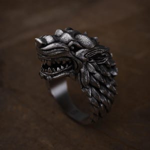 May include: A detailed silver ring featuring a snarling wolf head. The ring showcases intricate detailing of the wolf's fur, teeth, and facial features. The ring is a bold statement piece, perfect for those who appreciate animal-themed jewelry.