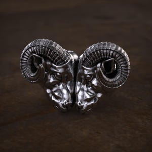May include: A silver-colored ring featuring a detailed design of two ram heads facing each other. The ram heads have large, curved horns and are set against a dark, textured background.