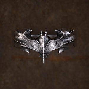 May include: A silver-colored ring featuring the iconic Batman symbol. The ring has a textured surface and a detailed bat-shaped emblem, with pointed wings and a central spike. The ring is likely made of metal.