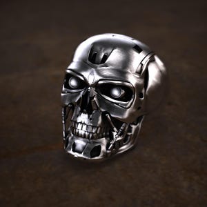 May include: A silver-colored skull ring with glowing white eyes. The ring has a detailed, metallic design, featuring a skeletal face with visible teeth and mechanical elements. The ring is likely made of metal and is a unique piece of jewelry.