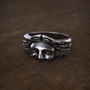 May include: A silver ring featuring a sculpted face with closed eyes and flowing hair. The ring has a detailed, textured design and a dark, antique finish. The face is centered on the ring, with the hair forming a frame around it.