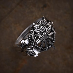 May include: A detailed silver ring featuring a central design with a stylized figure and a wheel-like element. The ring has intricate detailing and a polished finish, showcasing a gothic or fantasy theme.