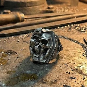 May include: A silver-colored ring with a skull design, featuring a half-face and skull motif. The ring is set on a metal workbench with tools, suggesting a handcrafted or artisan product. The ring's design is detailed and gothic.